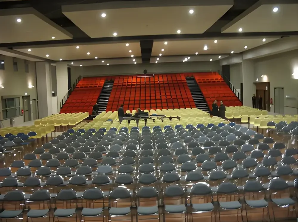 Salle de spectacle vide avec des fauteuils rouges et des chaises grises. La pièce est éclairée par des spots. L'ambiance générale est assez sombre et austère.
