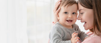 jeune femme brune aux cheveux longs qui porte dans ses bras un bébé de 6 mois environ très souriant.