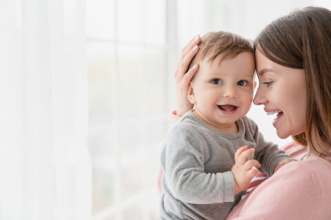 jeune femme brune aux cheveux longs qui porte dans ses bras un bébé de 6 mois environ très souriant.