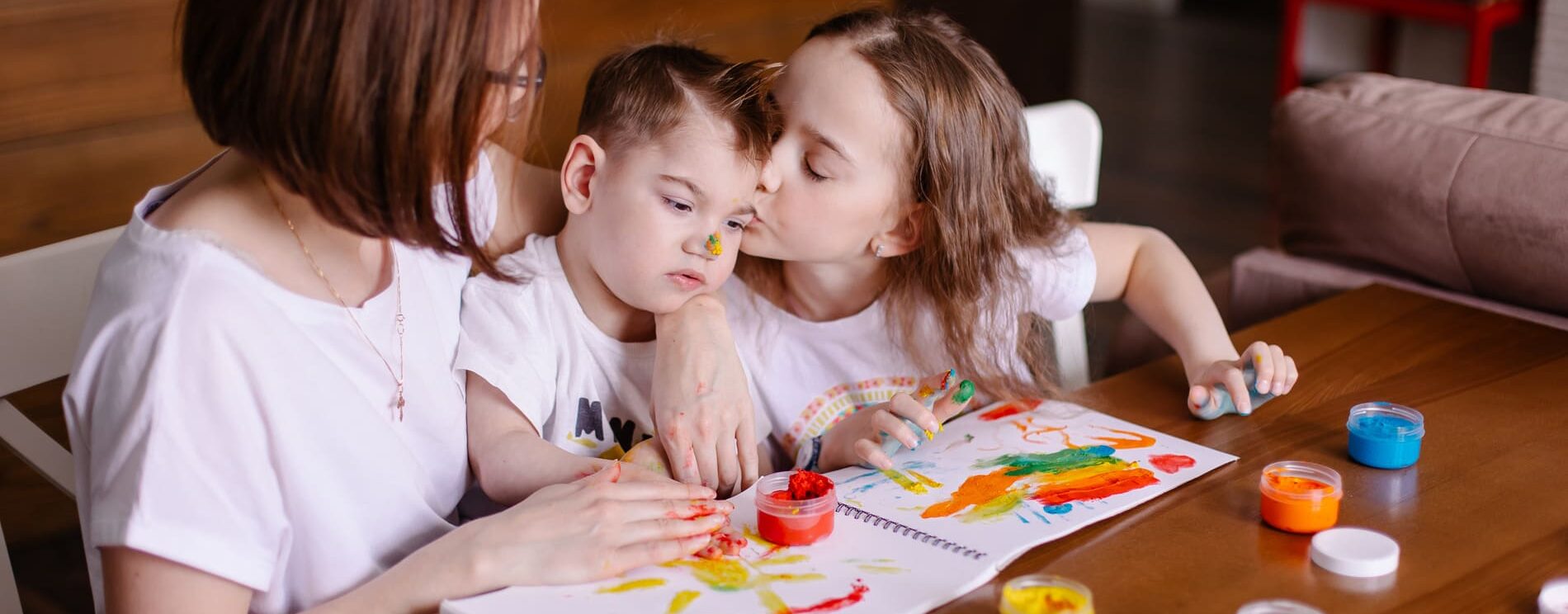 2 enfants avec leur maman ou une assistante maternelle. le petit garçon semble porteur d'un handicap. Les deux enfants font de la peinture sur un cahier posé sur la table devant eux.