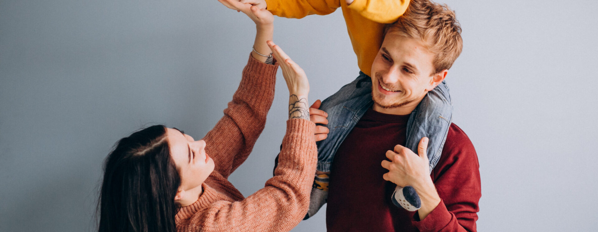 couple avec un enfant sur les épaules du père. L'enfant porte un pull jaune. le père un pull roiuge et un pantalon marron. La femme un pull rose orangé. La famille a l'air de s'amuser.