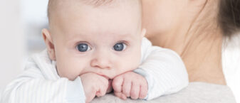 bébé aux grands yeux bleus porté par une femme brune. Le focus est fait sur le bébé uniquement. Il porte un pull blanc avec des rayures bleu clair et a les poings serrés sur l'épaule de la femme.