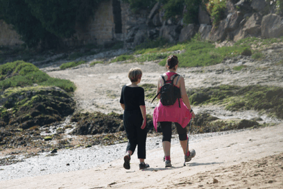 2 femmes, prises de dos, et habillées en tenue de sport ou de randonnée se promènent sur le littoral. Le sol est sableux et il y a un peu de végétation dans le fond de l'image. - Agrandir l'image, fenêtre modale