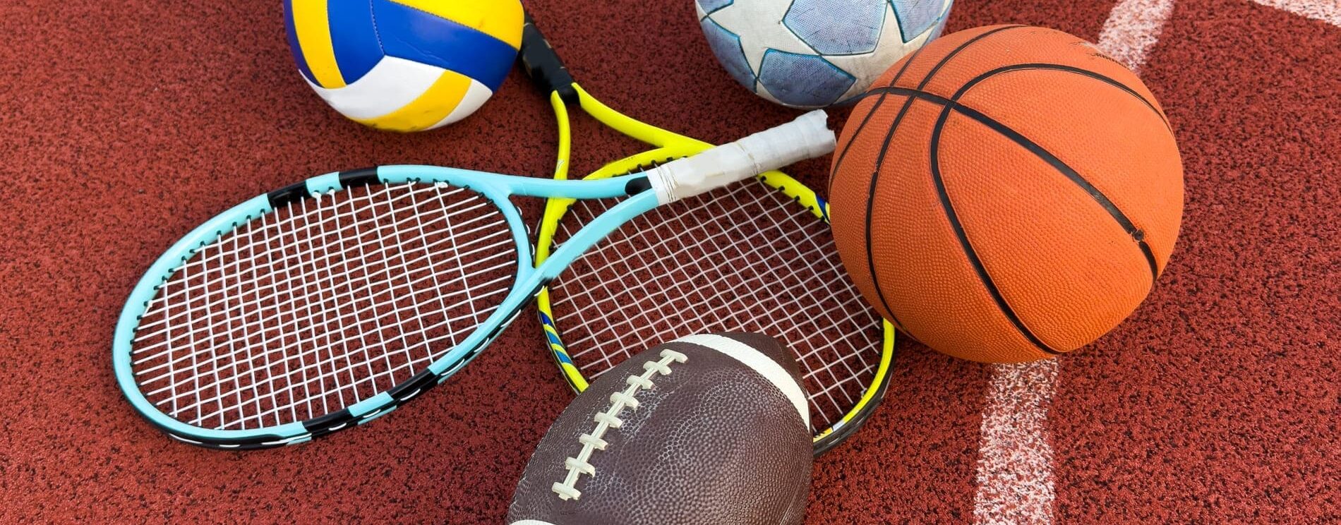 matériel de sport : ballons de football, de handball et de rugby, ainsi que raquettes de tennis posés sur la surface d'un terrain de sport