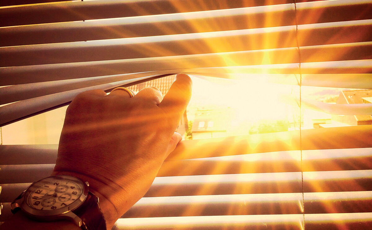 personne qui regarde l'extérieur au travers de persiennes. Grand soleil à l'extérieur. Pièce plongée dans l'obscurité