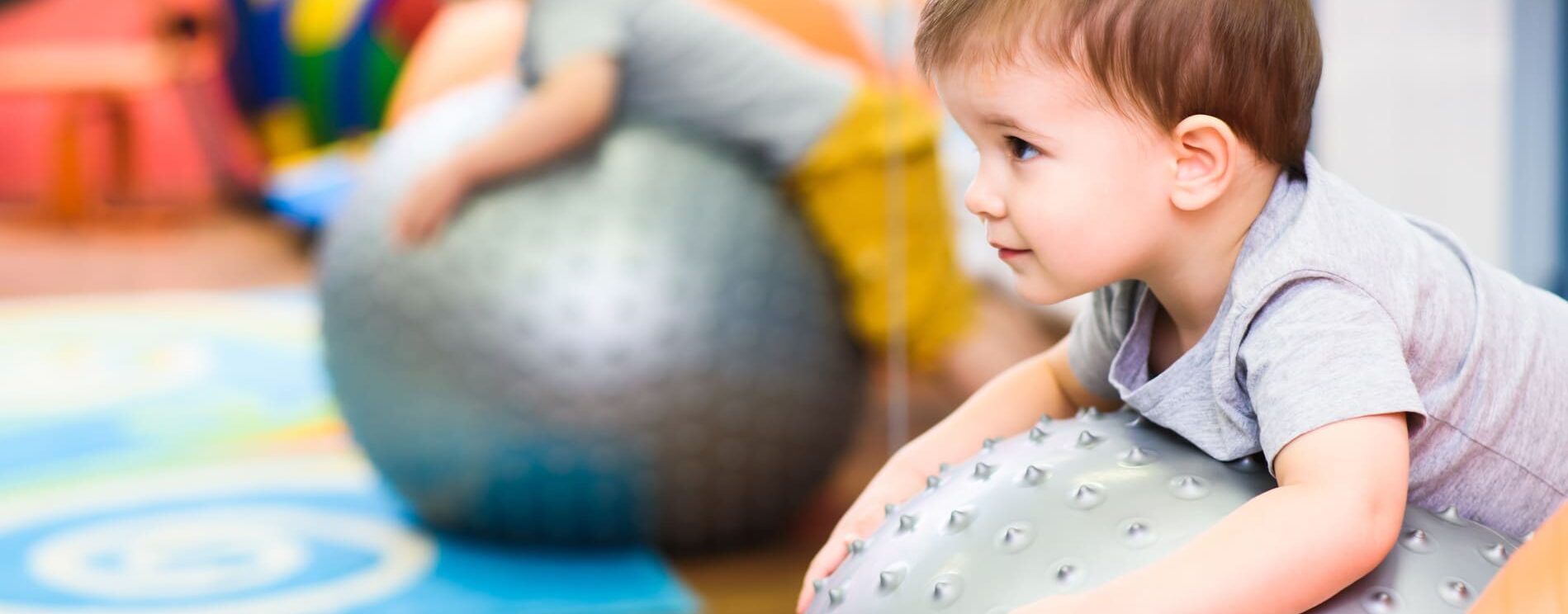 jeune enfant qui joue, allongé sur le ventre sur un gros ballon gris. environnement très coloré d'une crèche ou école maternelle