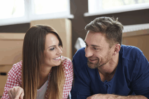 Jeune couple allongé par terre. Les deux personnes se regardent. Lui porte une chemise bleue et elle porte une chemise vichy rose et blanche. Ils sourient en se regardant.