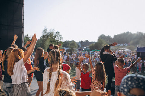 Groupe d'enfants pris de dos, sur scène, lors d'un concert en extérieur. Un musicien est sur scène et on devine la foule qui l'acclame.
