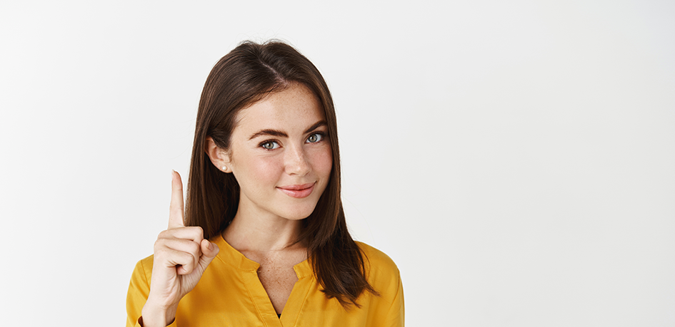 Jeune femme aux cheveux longs et bruns portant une chemise jaune. Elle lève le doit pour signifier de faire attention à quelque chose.