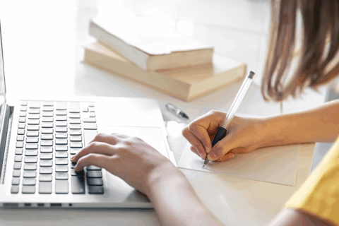 Femme aux cheveux mi-longs bruns qui écrit sur une page blanche avec un stylo. Elle a une main posée sur le clavier d'un ordinateur portable.