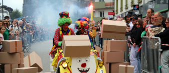 scène issue de la fête "du barouf dans l'bourg" à Guipavas. Un clown, en pleine rue, pousse une voiturette jaune et fait tomber des piles de cartons sur son passage. Nombreux spectateurs.