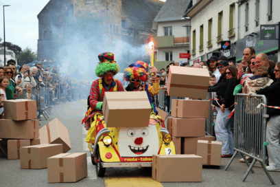 scène issue de la fête "du barouf dans l'bourg" à Guipavas. Un clown, en pleine rue, pousse une voiturette jaune et fait tomber des piles de cartons sur son passage. Nombreux spectateurs. - Agrandir l'image, fenêtre modale