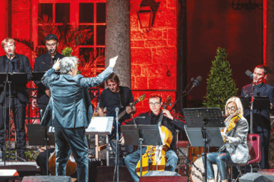 musiciens (violoncelle, guitare ou basse), flûte à bec et chanteur sur scène. Derrière eux, un mur en briques éclairés d'une lumière rouge. Devant eux, un chef d'orchestre qui lève les bras.