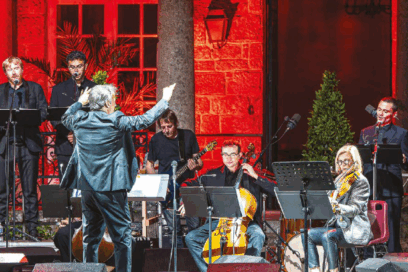 musiciens (violoncelle, guitare ou basse), flûte à bec et chanteur sur scène. Derrière eux, un mur en briques éclairés d'une lumière rouge. Devant eux, un chef d'orchestre qui lève les bras. - Agrandir l'image, fenêtre modale
