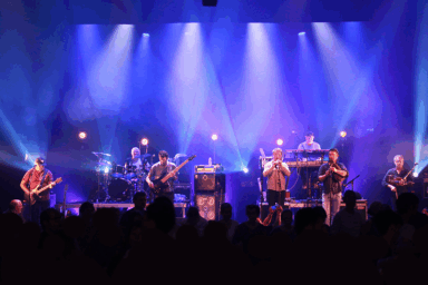 Musiciens qui jouent d'instruments issus de la culture bretonne (bombarde, cornemuse) et d'autres plus classiques (guitare ou basse). C'est un concert. Derrière eux, un homme joue du piano et un autre de la batterie. La cène est éclairée par des spots qui donnent une lumière bleutée à la scène.