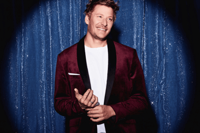 Jeremy Credeville. Homme blond costume noir sur scène, devant un rideau bleu argenté, souriant. - Agrandir l'image, fenêtre modale