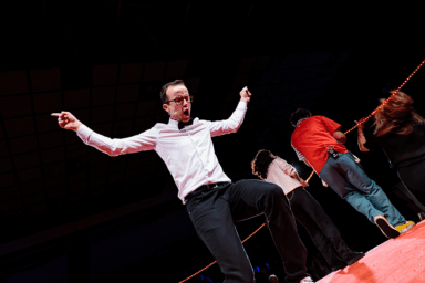 Spectacle d'improvisation. jeune homme sur scène, bras levés, qui semble parler très fort ou crier. En arrière-pla, deux personnes qui tournent le dos à l'objectif.