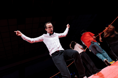 Spectacle d'improvisation. jeune homme sur scène, bras levés, qui semble parler très fort ou crier. En arrière-pla, deux personnes qui tournent le dos à l'objectif. - Agrandir l'image, fenêtre modale