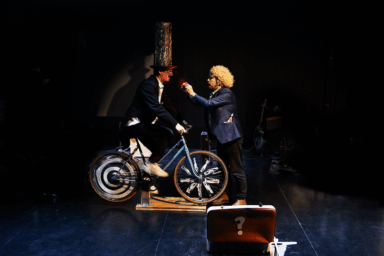 scène de théâtre issue de la pièce "mystèere et boule de gomme". 2 hommes sur scène, qui portent des vêtements qui s'apparentent à ceux de clowns : l'une d'entre eux est sur un vélo. L'autre porte une perruque blonde frisée et lui tend la main pour lui donner quelque chose.