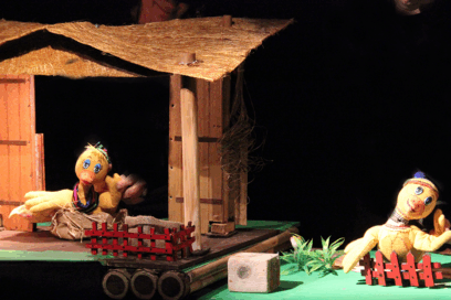 spectacle de marionnettes. 2 carnards sur un sol vert. L'un d'eux est devant une cabane en bois. L'autre placé derrière une petite barrière rouge. Le spectacle s'appelle "marionnettes en folie". - Agrandir l'image, fenêtre modale