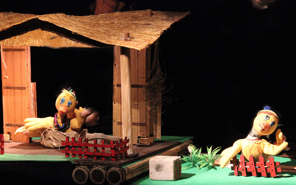 spectacle de marionnettes. 2 carnards sur un sol vert. L'un d'eux est devant une cabane en bois. L'autre placé derrière une petite barrière rouge. Le spectacle s'appelle "marionnettes en folie".