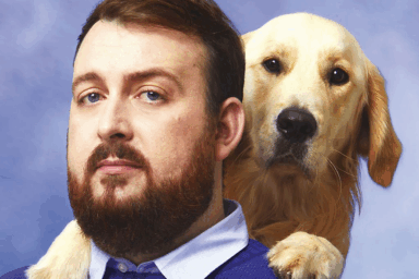 Artiste Moguiz. Un homme en gros plan. Pull-over bleu-violet, barbe rousse, cheveux courts, avec un chien de race labrador ou golden retriever sur l'paule.