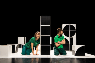 Pièce de théâtre "tout est chamboulé". Un homme et une femme sur une scène de théâtre. Fond noir. En décoration : de gros cubes noirs et blancs empilés de façon aléatoire. L'homme croise les bras et regarde vers l'extérieur. La femme est appuyée contre l'une des tours de cubes .