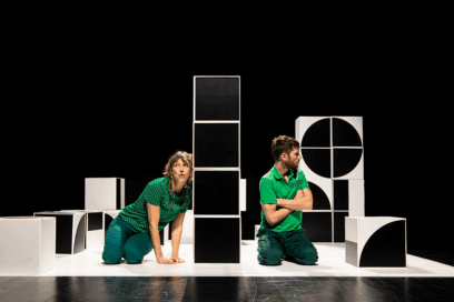 Pièce de théâtre "tout est chamboulé". Un homme et une femme sur une scène de théâtre. Fond noir. En décoration : de gros cubes noirs et blancs empilés de façon aléatoire. L'homme croise les bras et regarde vers l'extérieur. La femme est appuyée contre l'une des tours de cubes . - Agrandir l'image, fenêtre modale