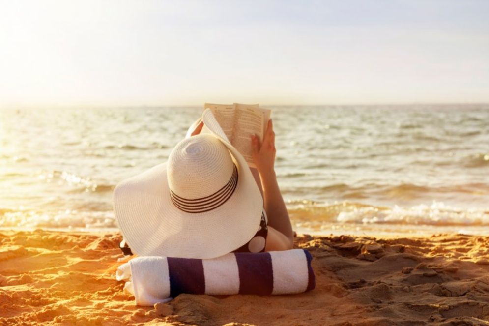 une femme de dos portant un grand chapeau blanc lisant un livre allongée sur une place, au soleil couchant devant la mer